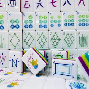 May include: A collection of white Mahjong tiles featuring colorful designs including numbers, bamboo patterns, dragons, and joker tiles with a crown graphic. The tiles have layered edges in purple, lime green, and blue, with some tiles standing upright.