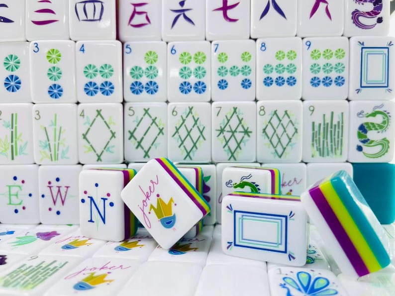 Puede incluir: Una colecci&oacute;n de fichas de Mahjong blancas con dise&ntilde;os coloridos que incluyen n&uacute;meros, patrones de bamb&uacute;, dragones y fichas de comod&iacute;n con un gr&aacute;fico de corona. Las fichas tienen bordes en capas de color morado, verde lima y azul.