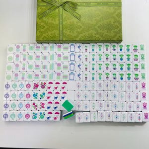 May include: A mahjong set featuring white tiles with colorful illustrations of flowers, bamboo, lanterns, and Chinese characters, arranged in rows. The set includes a green rectangular storage box with a repeating pattern and a green ribbon bow.