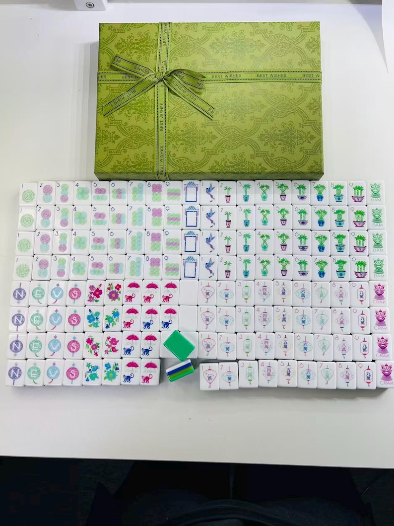 May include: A mahjong set featuring white tiles with colorful illustrations of flowers, bamboo, lanterns, and Chinese characters, arranged in rows. The set includes a green rectangular storage box with a repeating pattern and a green ribbon bow.
