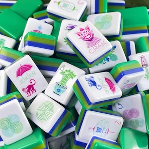 May include: A collection of rectangular Mahjong tiles featuring white faces with colorful illustrations, including a pink umbrella, a monkey, a green plant, and the text JOKER and CRAK, with layered green, blue, and white sides.