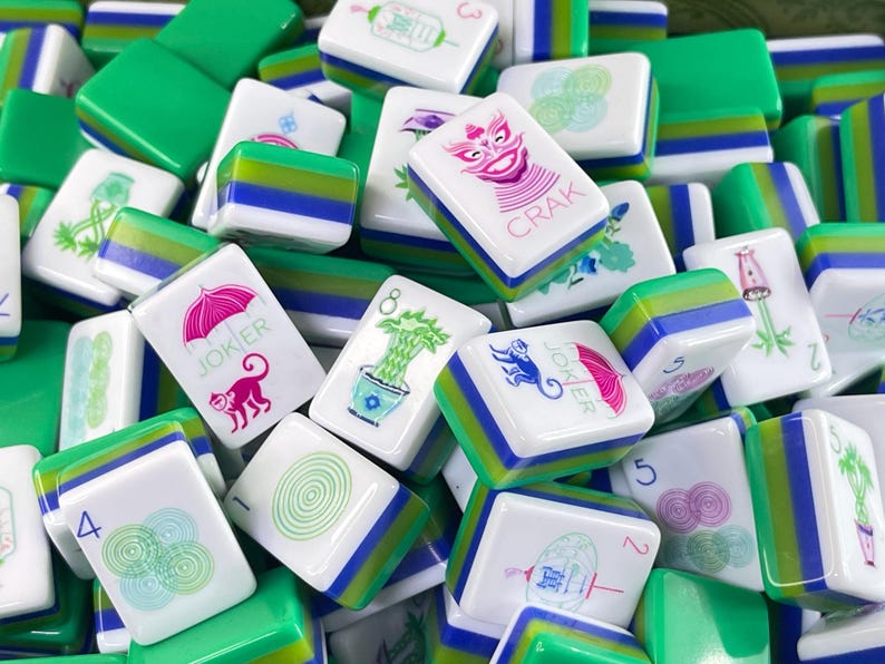 May include: A collection of rectangular Mahjong tiles featuring white faces with colorful illustrations, including a pink umbrella, a monkey, a green plant, and the text JOKER and CRAK, with layered green, blue, and white sides.