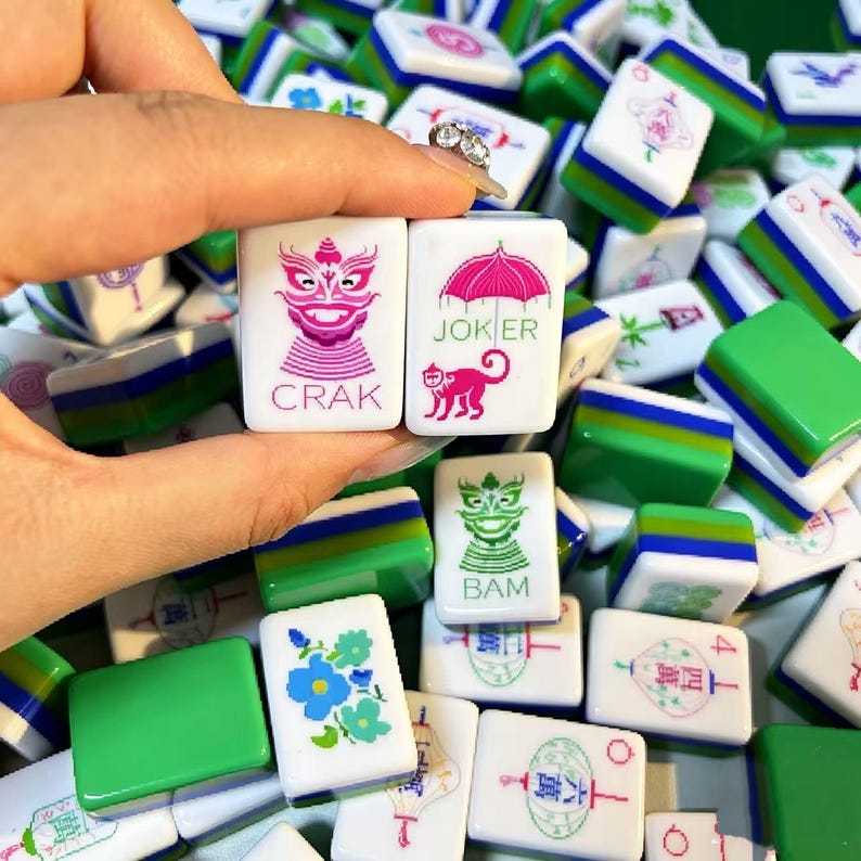 May include: A collection of white and green Mahjong tiles featuring various colorful engraved designs. Two tiles are held up, one displaying a pink dragon with the text CRAK and another showing a pink monkey under an umbrella with the text JOKER.