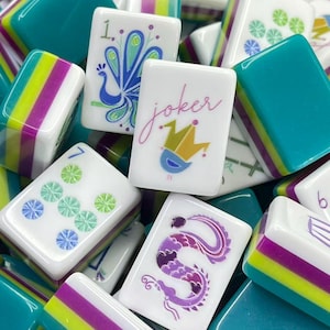 Puede incluir: Una colecci&oacute;n de fichas de Mahjong con caras blancas e ilustraciones coloridas que incluyen un pavo real, un comod&iacute;n con corona, un drag&oacute;n y patrones num&eacute;ricos. Las fichas tienen lados con rayas horizontales en color verde azulado, verde lima y morado.