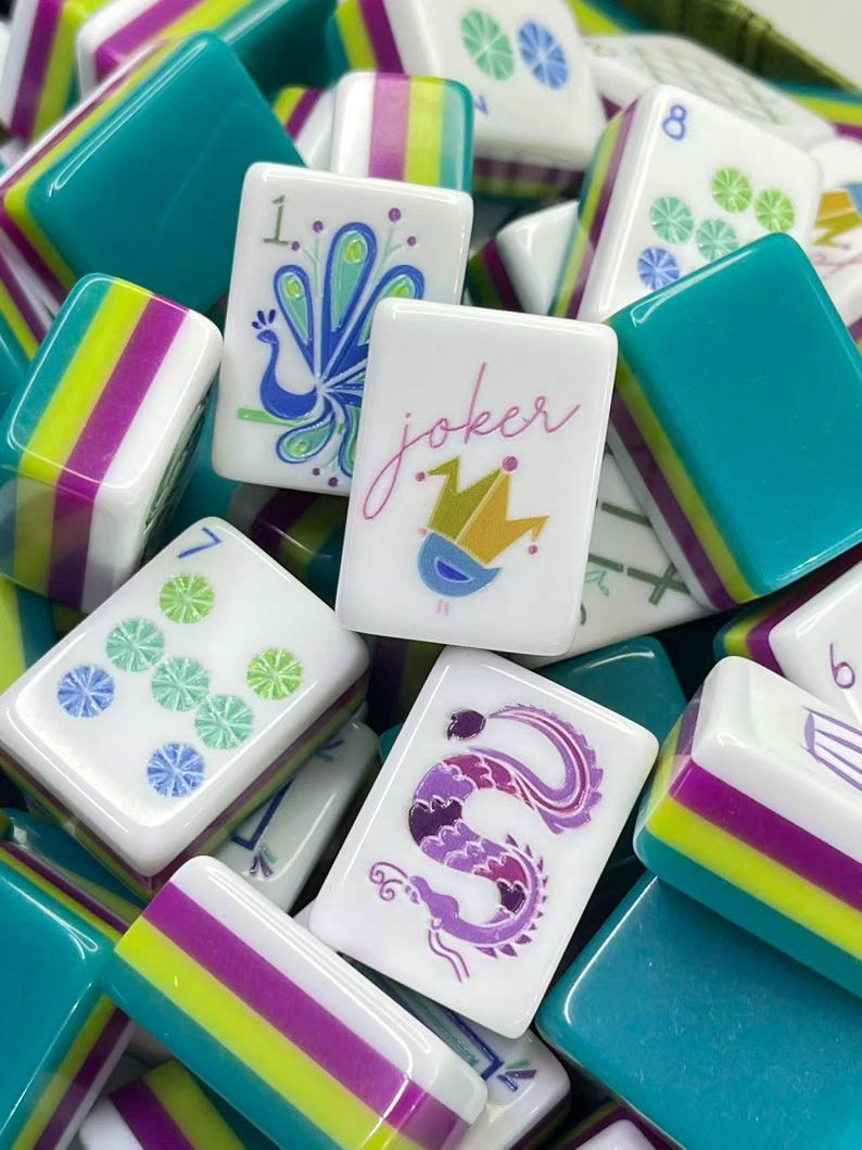 Puede incluir: Una colecci&oacute;n de fichas de Mahjong con caras blancas e ilustraciones coloridas que incluyen un pavo real, un comod&iacute;n con corona, un drag&oacute;n y patrones num&eacute;ricos. Las fichas tienen lados con rayas horizontales en color verde azulado, verde lima y morado.