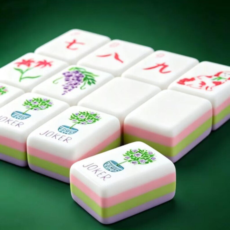 May include: A set of rectangular mahjong tiles with white tops featuring colorful illustrations of flowers, plants, and the word JOKER. Each tile has a layered base with horizontal stripes in pink, light green, and light purple colors.