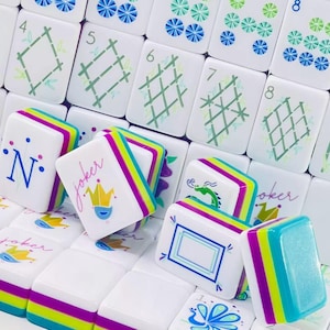 Puede incluir: Una colecci&oacute;n de fichas de Mahjong blancas con dise&ntilde;os coloridos que incluyen tallos de bamb&uacute;, rejillas geom&eacute;tricas, n&uacute;meros y la palabra joker con un icono de corona. Las fichas tienen bordes en capas en tonos brillantes de morado, verde lima y azul.