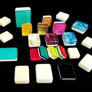 May include: A collection of rectangular mahjong tiles featuring various finishes including solid white, translucent teal, glittery pink, gold foil, marbled blue, and multi-colored striped designs. The tiles are arranged in a scattered formation.