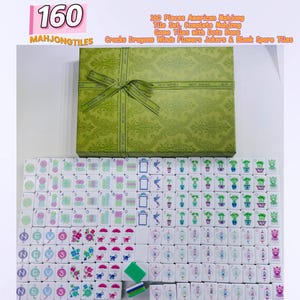 May include: A 160-piece American Mahjong tile set with a green patterned storage box featuring a ribbon. The white tiles display various colorful symbols including dots, bams, cracks, dragons, winds, flowers, jokers, and blank spare tiles.