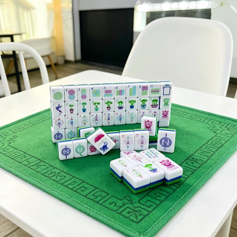 May include: A set of white Mahjong tiles with colorful illustrations of plants, lanterns, and animals, arranged in rows and stacks on a green felt mat. The tiles feature numbers and symbols in blue, green, pink, and purple, with some tiles showing a cat.