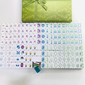 Puede incluir: Un juego de mahjong con fichas blancas que presentan s&iacute;mbolos, n&uacute;meros y dise&ntilde;os florales grabados en color, acompa&ntilde;ado de una caja de regalo verde claro con una cinta a juego impresa con las palabras BEST WISHES.