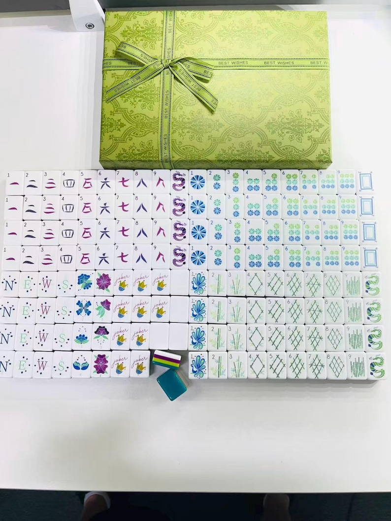Puede incluir: Un juego de mahjong con fichas blancas que presentan s&iacute;mbolos, n&uacute;meros y dise&ntilde;os florales grabados en color, acompa&ntilde;ado de una caja de regalo verde claro con una cinta a juego impresa con las palabras BEST WISHES.