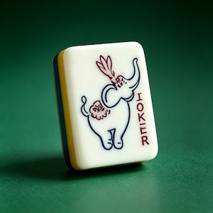 Op de afbeelding: Een rechthoekige mahjongtegel met een crèmekleurige voorzijde met een donkerblauwe omtrek van een olifant met roze bloemaccenten en het woord JOKER in het rood gedrukt. De zijkant van de tegel toont lagen van geel en donkerblauw kunststof.