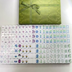 Puede incluir: Un juego de Mahjong con fichas blancas que presentan diversos s&iacute;mbolos, n&uacute;meros y caracteres de colores, dispuestos en filas. El conjunto incluye una caja de regalo rectangular verde con un patr&oacute;n de damasco y un lazo en la parte superior.