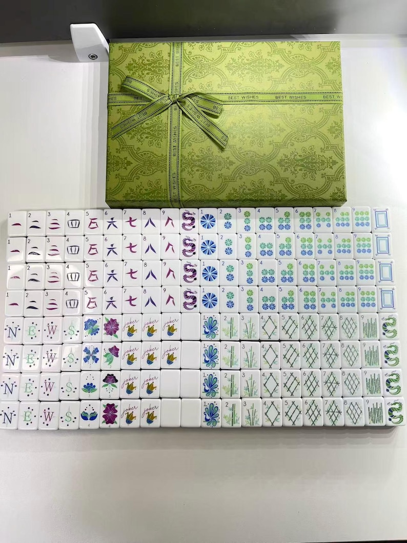 Puede incluir: Un juego de Mahjong con fichas blancas que presentan diversos s&iacute;mbolos, n&uacute;meros y caracteres de colores, dispuestos en filas. El conjunto incluye una caja de regalo rectangular verde con un patr&oacute;n de damasco y un lazo en la parte superior.