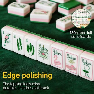 May include: A set of white mahjong tiles with pink and green accents, featuring numbered tiles from one to three, palm leaf illustrations, and tiles labeled Joker. The set includes 160 pieces with polished edges, displayed on a dark green surface.