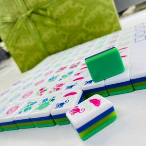 May include: A set of white mahjong tiles with green, blue, and white layered bases. The tiles feature colorful printed graphics including pink umbrellas, blue monkeys, and floral patterns. One tile displays the word JOKER in pink text.