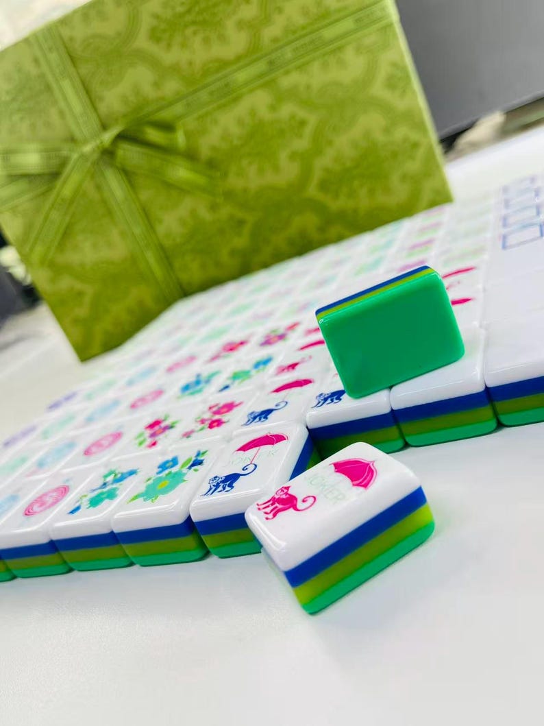 May include: A set of white mahjong tiles with green, blue, and white layered bases. The tiles feature colorful printed graphics including pink umbrellas, blue monkeys, and floral patterns. One tile displays the word JOKER in pink text.