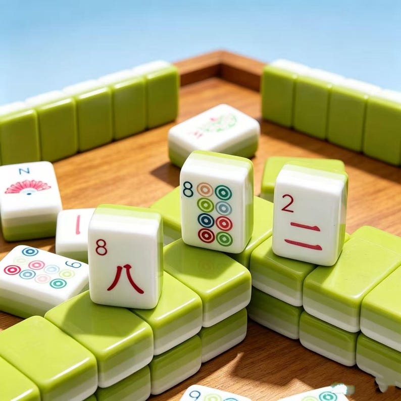 May include: A set of rectangular mahjong tiles with white faces and light green backs. The tiles feature various red, green, and blue symbols, including numbers and circular patterns, arranged on a wooden surface with some tiles standing upright.