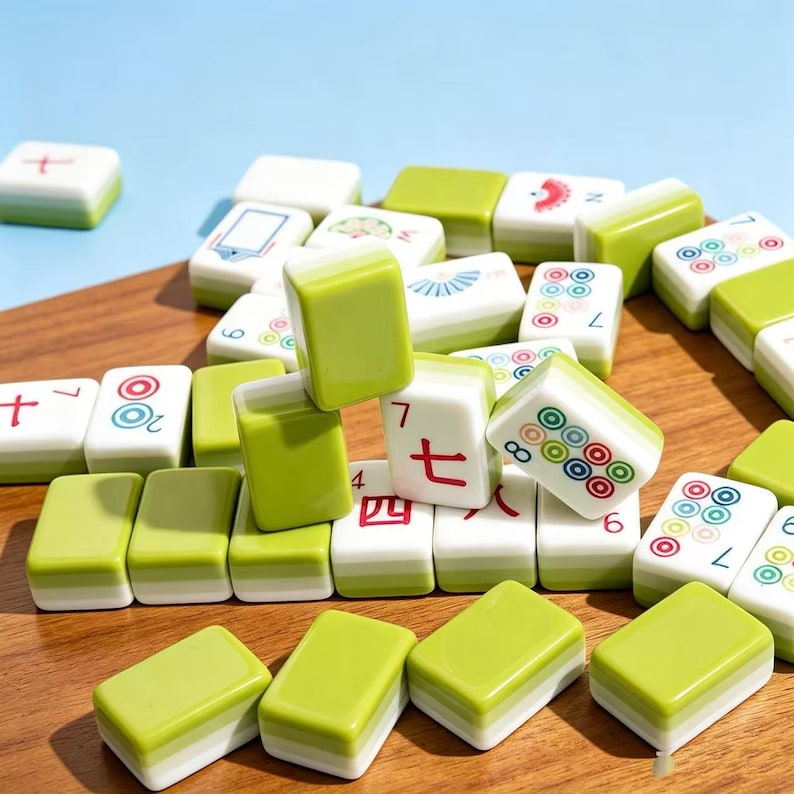 May include: A collection of rectangular mahjong tiles with a two-tone design featuring a light green top and a white base. The tiles display various colorful symbols, Chinese characters, and circular patterns, scattered across a wooden surface.