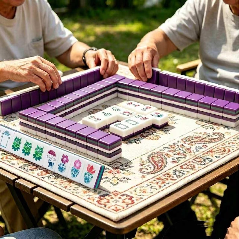 Pu&ograve; includere: Un set di tessere da mahjong viola e bianche disposte su un tappetino in tessuto fantasia. Le tessere presentano vari disegni floreali e a ombrello verdi e rosa sulle facce. I pezzi del gioco sono organizzati in una formazione rettangolare.