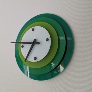 May include: A circular wall clock featuring three layered discs in shades of dark green, lime green, and white. The clock has black hands and four small black circular markers on the white face, creating a tiered, multi-dimensional appearance.