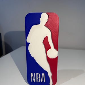 Puede incluir: Logotipo de la NBA impreso en 3D en azul, rojo y blanco. Presenta la silueta de un jugador de baloncesto botando el balón, con las letras "NBA" en la parte inferior. El logotipo es rectangular con esquinas redondeadas.