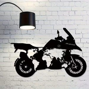 Peut inclure: Décoration murale en métal noir représentant une moto avec une carte du monde intégrée. La silhouette de la moto porte l'inscription "R1200". L'œuvre est fixée sur un mur de briques blanches. Une lampe noire est suspendue au-dessus.
