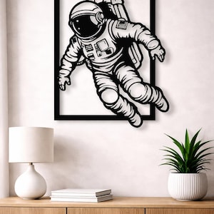 Peut inclure: Décoration murale en métal noir et blanc représentant un astronaute flottant dans l'espace. L'astronaute porte une combinaison spatiale et un casque, encadré dans un rectangle noir. Sur un mur blanc, au-dessus d'un meuble en bois.