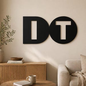Peut inclure: Décoration murale en métal noir avec le mot "DO". Le "D" est une forme stylisée avec une découpe verticale, et le "T" est à l'intérieur d'un cercle. L'art est accroché sur un mur de couleur neutre dans un salon.