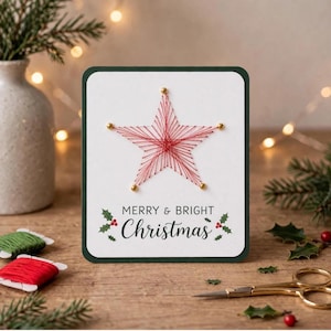 May include: A rectangular greeting card with a dark green border on a white background. The card features a red string art star design anchored by gold pins. Below the star, the text reads MERRY & BRIGHT Christmas in black, with holly.
