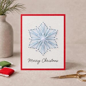 Puede incluir: Una tarjeta de felicitación rectangular con borde rojo y centro blanco que presenta un diseño de copo de nieve hecho con arte de hilos en color azul claro y blanco. El texto Merry Christmas está impreso en cursiva negra abajo.