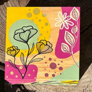 Puede incluir: Una pieza de arte cuadrada de madera con un diseño floral. La obra de arte presenta un fondo amarillo con un círculo verde y formas rosas y verdes. Flores negras y amarillas y hojas blancas están pintadas sobre la madera.