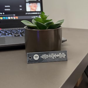 Può includere: Una piccola pianta succulenta verde in un vaso di ceramica marrone è su una scrivania accanto a un laptop. Un'etichetta rettangolare grigia con un codice Spotify è di fronte alla pianta. L'etichetta Spotify ha un logo Spotify bianco.