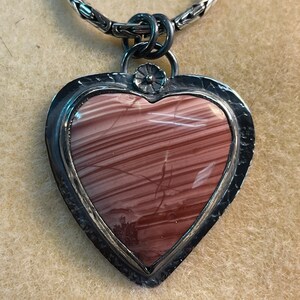 May include: A silver heart-shaped pendant with a brown and pink striped stone set in the center. The pendant is attached to a silver chain.