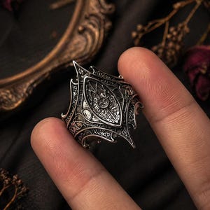 Peut inclure: Une bague en argent détaillée avec un design gothique orné. La bague présente un élément central en forme de bouclier avec des motifs complexes et des bords pointus. La bague est tenue entre deux doigts, mettant en valeur son savoir-faire.