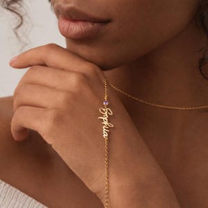 Peut inclure: Un bracelet doré avec le nom "Sophia" écrit en cursive. Le bracelet est orné d'une petite pierre précieuse violette. Un collier en or est également visible.