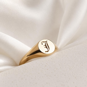 Peut inclure: Une bague chevalière dorée avec une finition polie. La bague présente une face circulaire avec la lettre "J" gravée dans une police classique. La bague est présentée sur un tissu blanc et soyeux.
