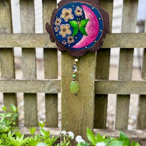May include: A wooden circle with a painted design of a pink crescent moon, green butterfly, and yellow flowers on a blue background. The circle is hanging from a wooden fence with a green and brown beaded string.