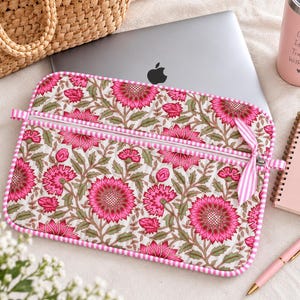 Puede incluir: Una funda acolchada rectangular para portátil con un vibrante estampado floral de flores de color rosa brillante y hojas verdes sobre un fondo crema. La funda tiene una cremallera blanca con un tirador de cinta a rayas rosas y blancas.