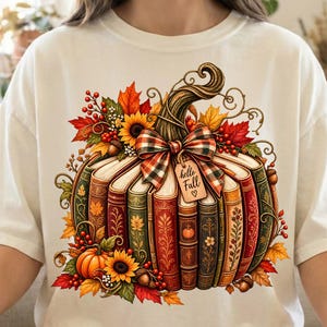 May include: A cream-colored t-shirt featuring a graphic of a pumpkin constructed from stacked vintage books. The design includes a plaid bow, a tag reading "hello Fall", sunflowers, red berries, and autumn leaves in shades of orange and red.