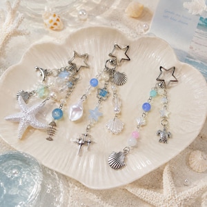 May include: Three silver-toned keychains featuring star-shaped clasps and dangling charms. The charms include white starfish, seashells, blue and pink beads, a fish skeleton, a four-pointed star, and a sea turtle, all arranged on silver chains.