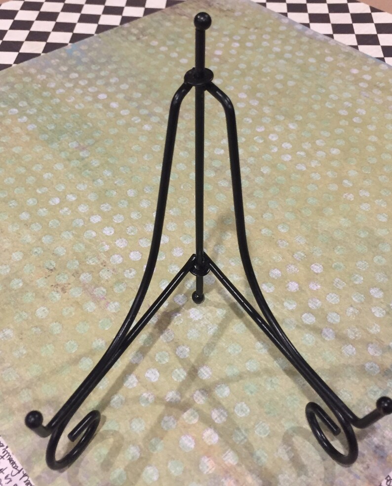May include: Black metal display stand with a triangular base and a curved top. The stand is designed to hold a plate or other flat object.