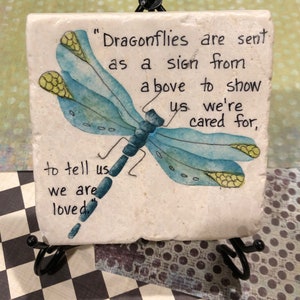 May include: A white ceramic coaster with a blue dragonfly painted on it. The coaster has the text "Dragonflies are sent as a sign from above to show us we're cared for, to tell us we are loved."