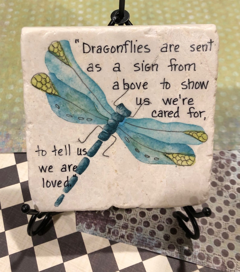 May include: A white ceramic coaster with a blue dragonfly painted on it. The coaster has the text "Dragonflies are sent as a sign from above to show us we're cared for, to tell us we are loved."