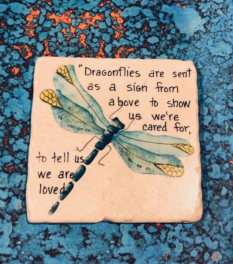May include: A white ceramic coaster with a blue dragonfly painted on it. The coaster has the text "Dragonflies are sent as a sign from above to show us we're cared for, to tell us we are loved".