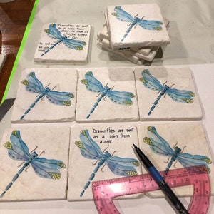 May include: Six square white coasters with blue and yellow dragonfly designs. The coasters have the text "Dragonflies are sent as a sign from above to show us we're cared for." and "Dragonflies are sent as a sign from above".