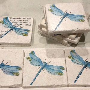 May include: Set of six square coasters with a white background and a blue dragonfly painted on each. The coasters have a quote about dragonflies being a sign from above to show us we are cared for.