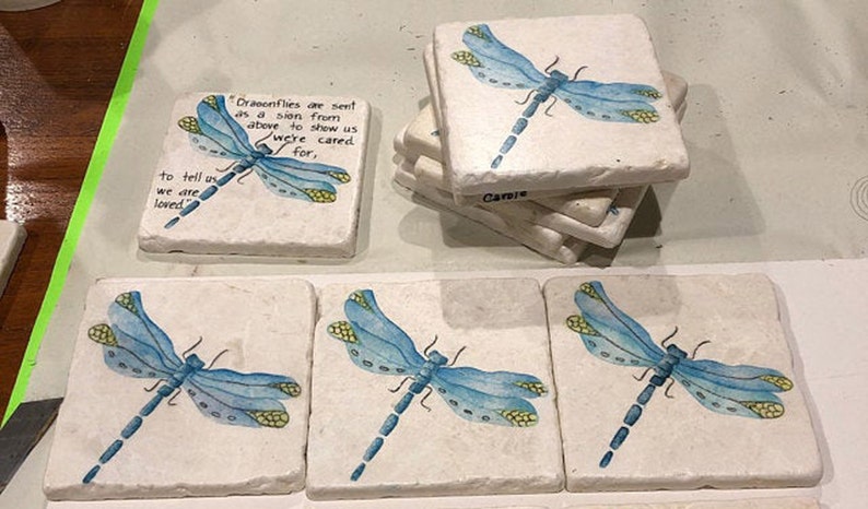 May include: Set of six square coasters with a white background and a blue dragonfly painted on each. The coasters have a quote about dragonflies being a sign from above to show us we are cared for.