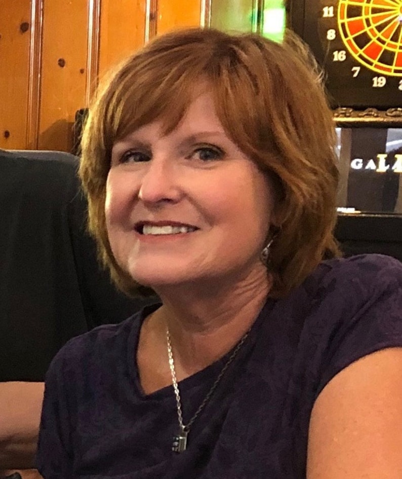May include: A woman with short, wavy, auburn hair is smiling. She is wearing a dark purple t-shirt with a silver chain necklace and a small pendant.